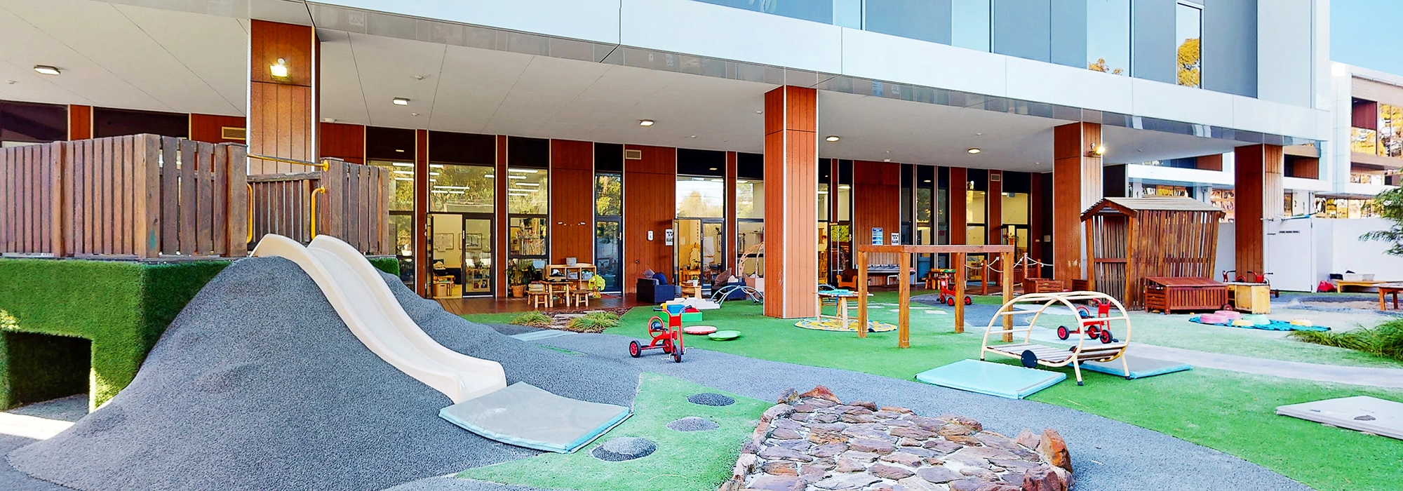 Outdoor playground area in front of a modern building with various play equipment like slides, tricycles, climbing frames, and a small wooden house on artificial grass and pebble surfacing.