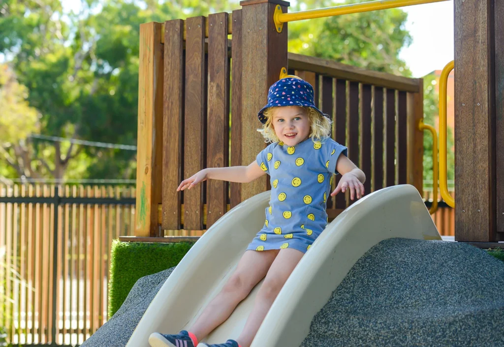 A young child wearing a hat and a T-shirt dress with smiley faces slides down a playground slide on a sunny day.