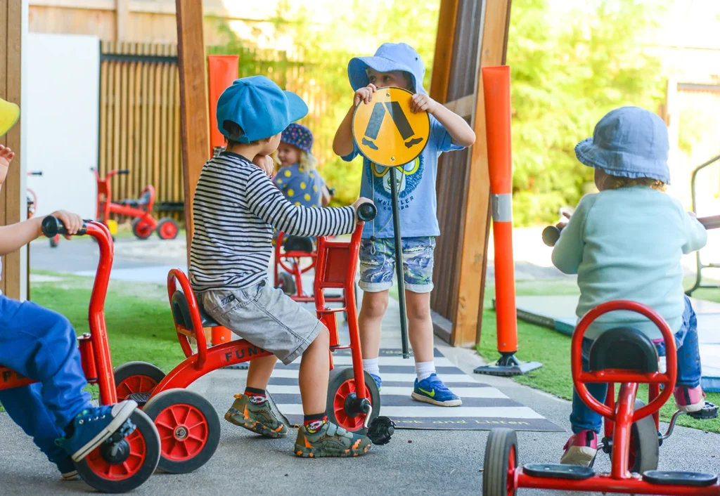 Several children play outdoors with tricycles and traffic signs in a play area. They wear hats for sun protection, and one child holds a caution sign.