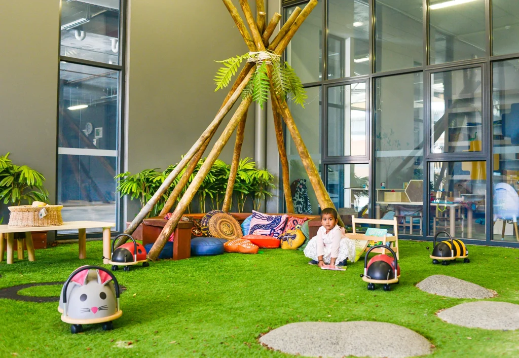 A child sits in a playroom with a tipi structure, cushions, and ride-on toys shaped like insects on a green carpet. Glass walls and indoor plants are in the background.