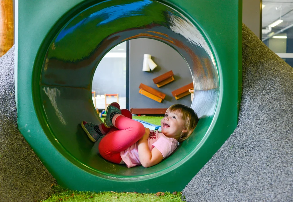 A young child wearing a pink outfit is sitting inside a large green playground tunnel, smiling and looking up. Various playground equipment is visible in the background.