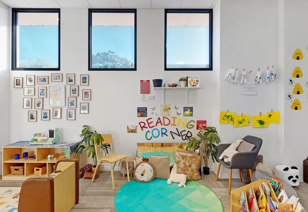 A bright, well-organized children's reading corner with a colorful rug, small chairs, shelves of books, potted plants, and cheerful wall decorations under two large windows.