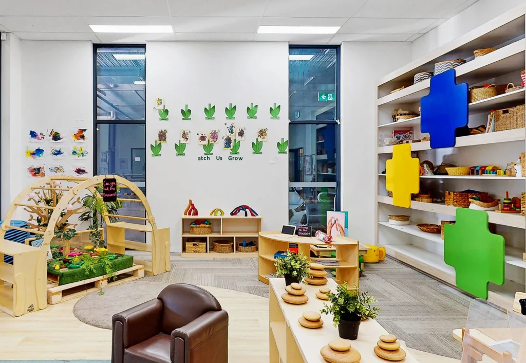 A colorful classroom with educational toys, bookshelves, green plants, and wall decorations. There is a brown armchair, a play arch, and a wall display reading "Watch Us Grow.