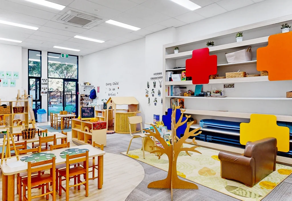 A bright, well-organized classroom with tables, chairs, bookshelves, and colorful wall decorations. There are various learning materials and toys arranged around the room.