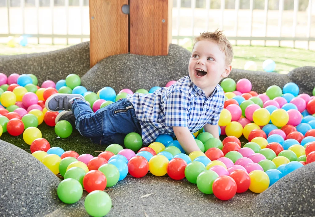 A young child in a blue checkered shirt and jeans is lying in a colorful ball pit, smiling and looking up. The background includes a fence and a wooden structure.