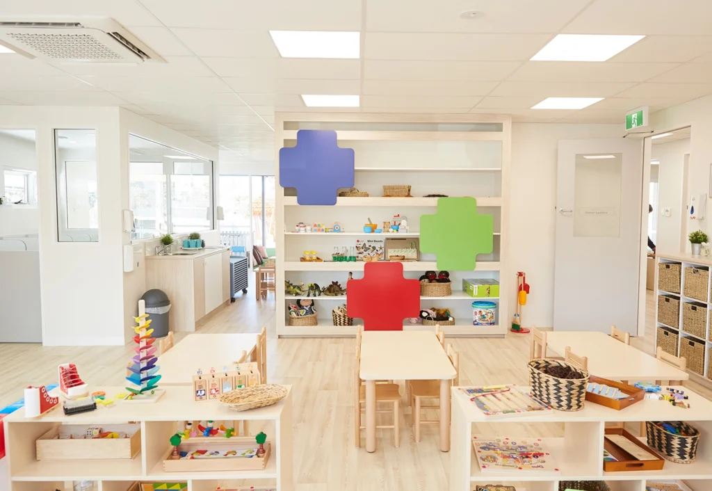 A brightly lit, clean preschool classroom with wooden furniture, shelves filled with toys, and colorful puzzle piece wall decorations.