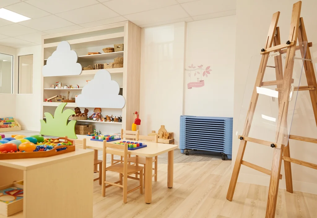 A brightly lit, organized classroom with shelves of toys, small tables and chairs, an easel, and a stack of blue floor mats against the wall.