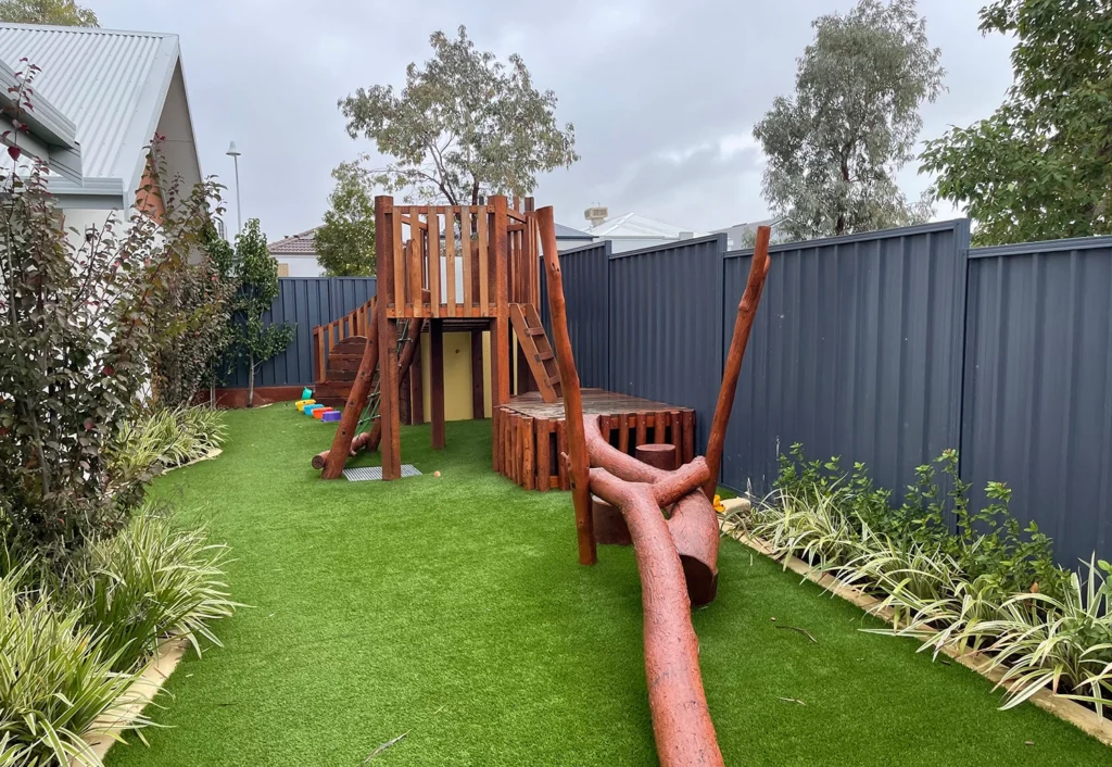 A backyard with artificial grass, featuring a wooden play structure with a platform, slide, and climbing elements. There are plants along the fence and a house to the left.