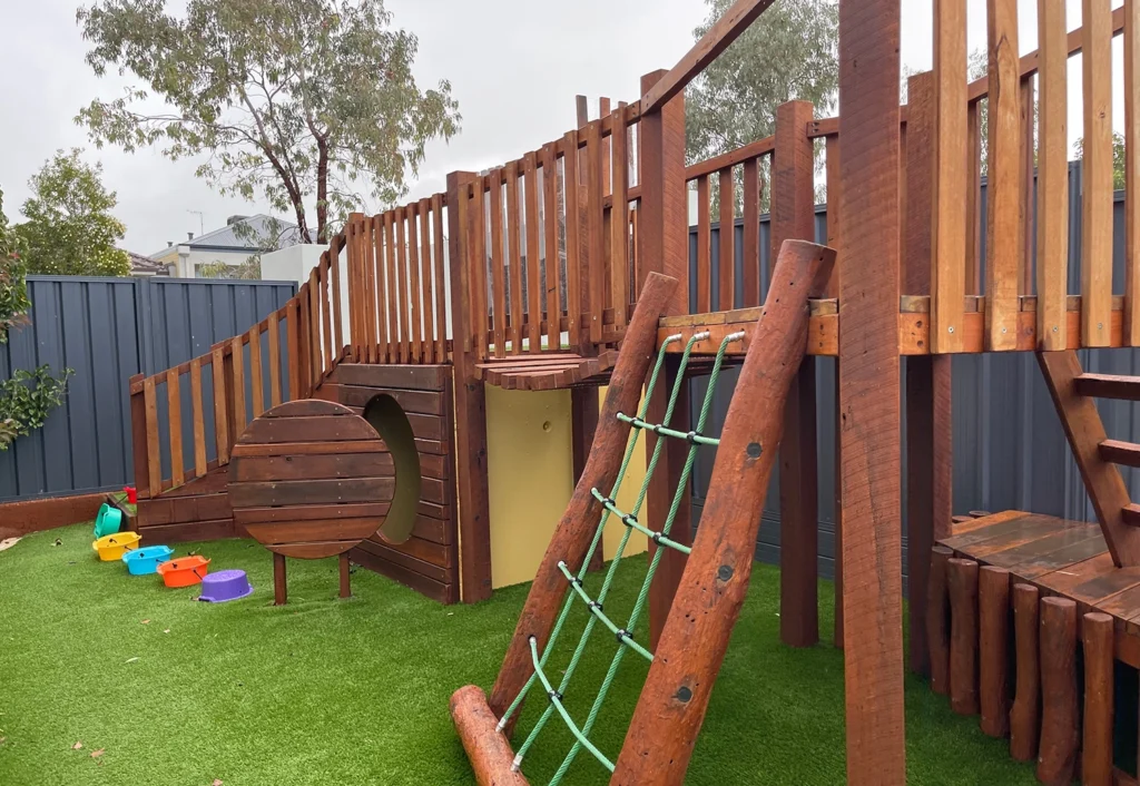 A children's outdoor play area with a wooden climbing frame, a green rope ladder, a tunnel, and colorful buckets on artificial grass. Trees and a blue fence are in the background.