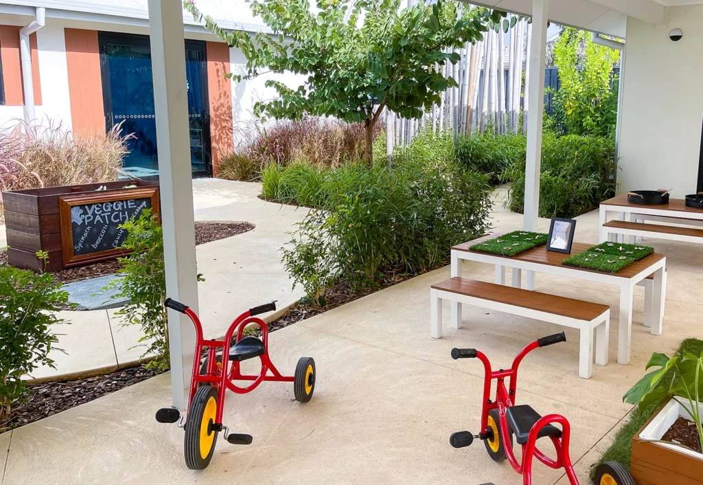 Outdoor play area with two red tricycles, a wooden table, and two benches. A planter labeled "Veggie Patch" is visible in the background, surrounded by greenery and a paved pathway.