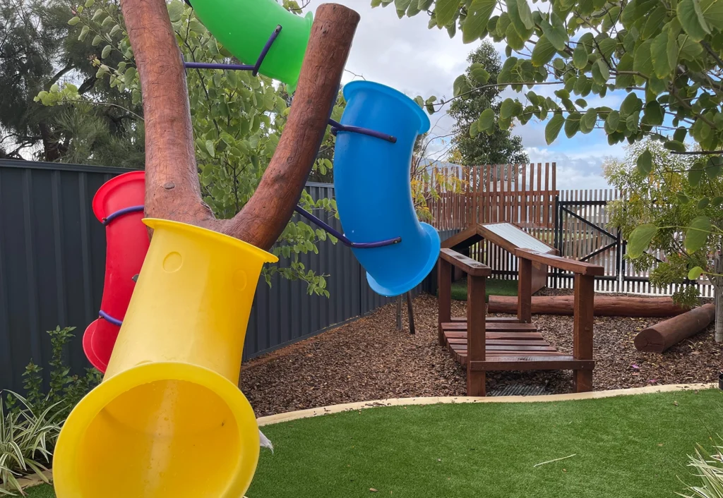 Colorful tube slides and a small wooden bridge in a children's outdoor playground area with surrounding greenery and a fenced background.