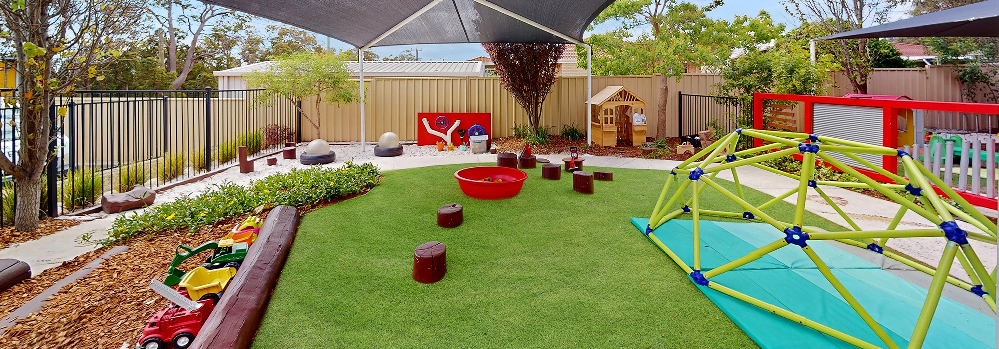 A children's outdoor play area at Byford childcare with various toys, green artificial turf, a climbing frame, wooden stepping stones, a sandpit, and a shade structure overhead.