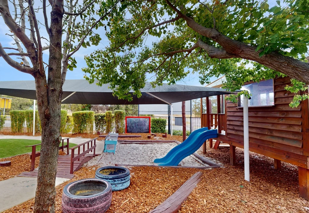 Outdoor playground at Busy Bees Byford North with a wooden cubby house, a blue slide, sandpit and shaded areas. Trees provide additional shade. A chalkboard is visible in the background.