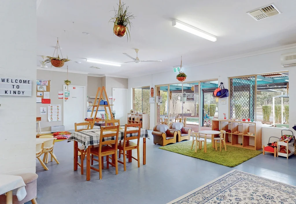 A brightly lit kindergarten classroom with wooden furniture, children's toys, craft supplies, plants hanging from the ceiling, and a sign reading "Welcome to Kindy".