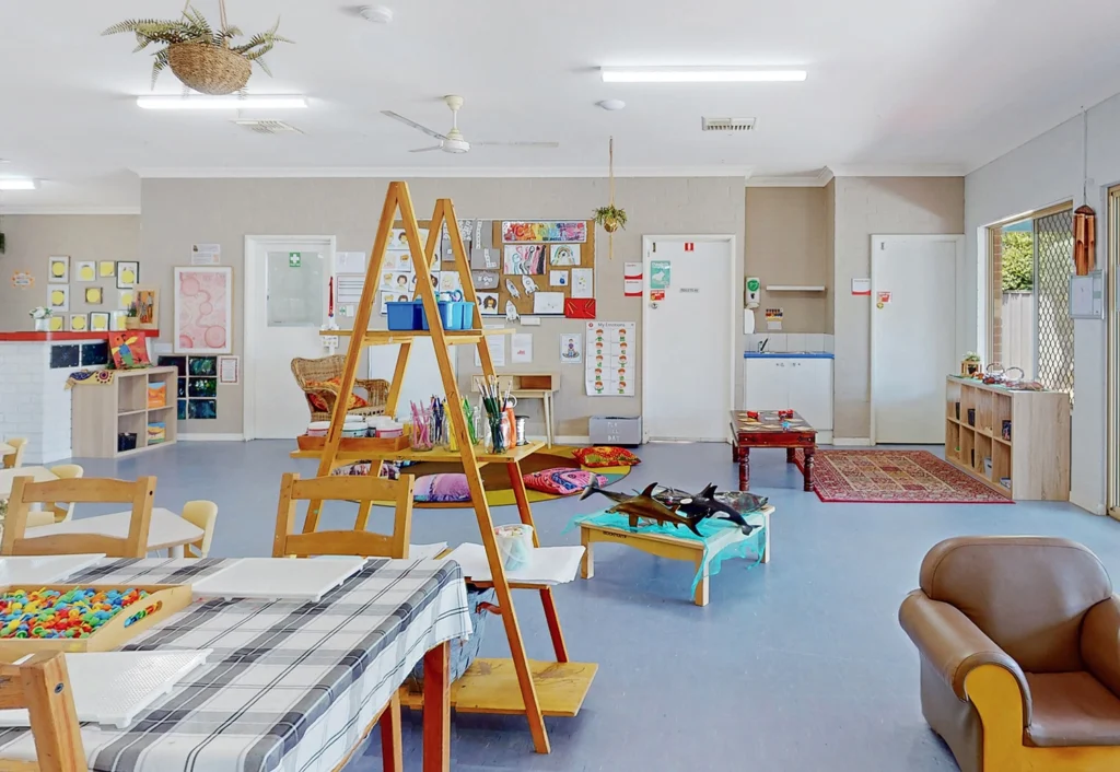 A brightly lit classroom at Byford childcare centre with art supplies, tables, chairs, an easel, and children's play areas. Walls are decorated with colorful artwork, and the room has a cozy and organized atmosphere.