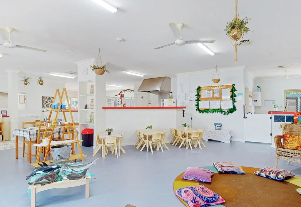 Brightly lit kindergarten classroom at Byford Child Care with small tables, chairs, hanging plants, and colorful decorations. Cushions are scattered on the floor, and an art station with supplies is set up in the corner.