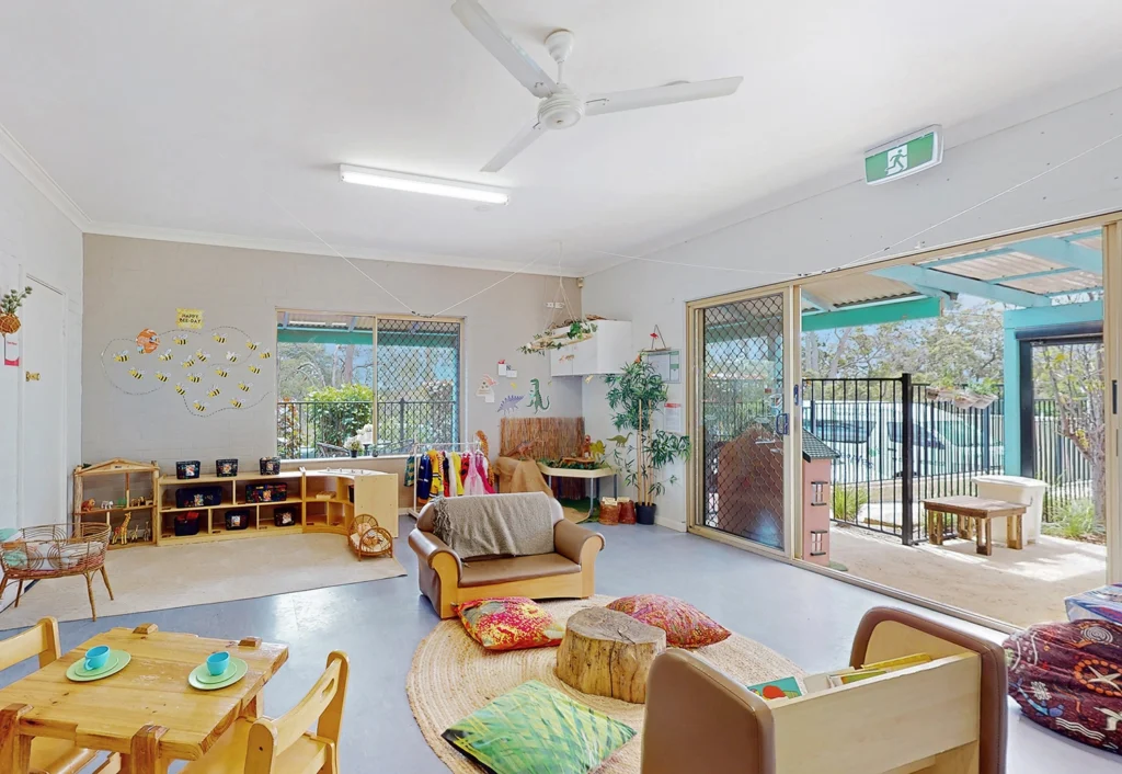 An indoor child care playroom with tables, chairs, toys, a couch, and a bookshelf. The space is bright with large windows and a ceiling fan. The room leads to an outdoor area through glass sliding doors.