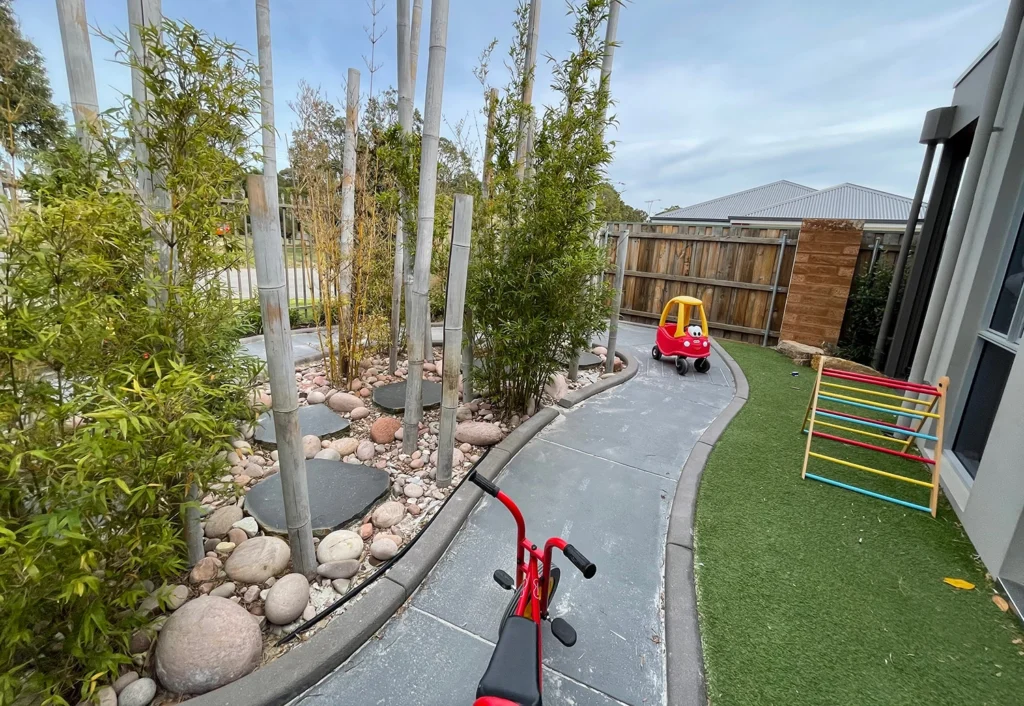 A red and yellow toy car, and a tricycle on a bike path at Byford child care centre. The middle of the bike track has bamboo plants, rocks and stepping stones for adventure.