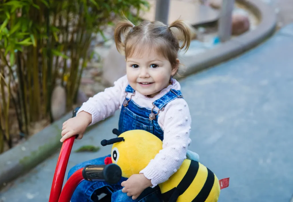 A young child with pigtails, wearing a denim overall and a pink sweater, holds a plush bee toy while sitting on a red tricycle at Byford daycare.