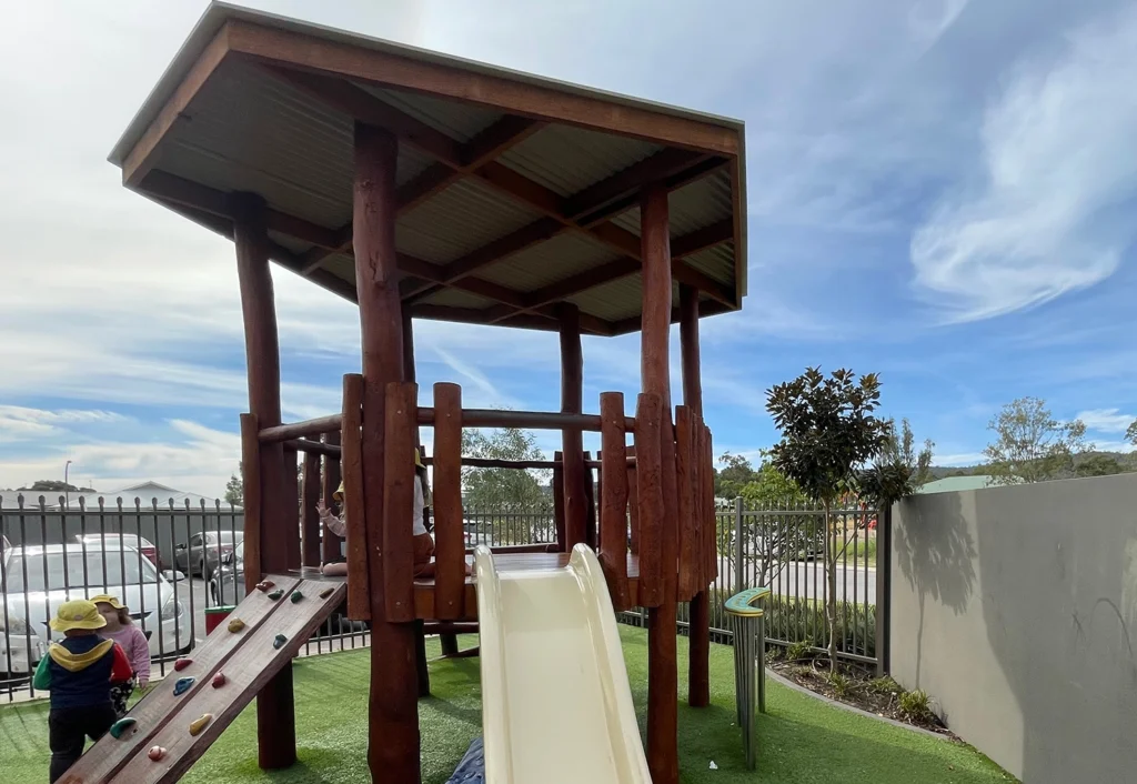 A shaded wooden playhouse with a slide and rock climbing wall at Busy Bees Byford childcare.