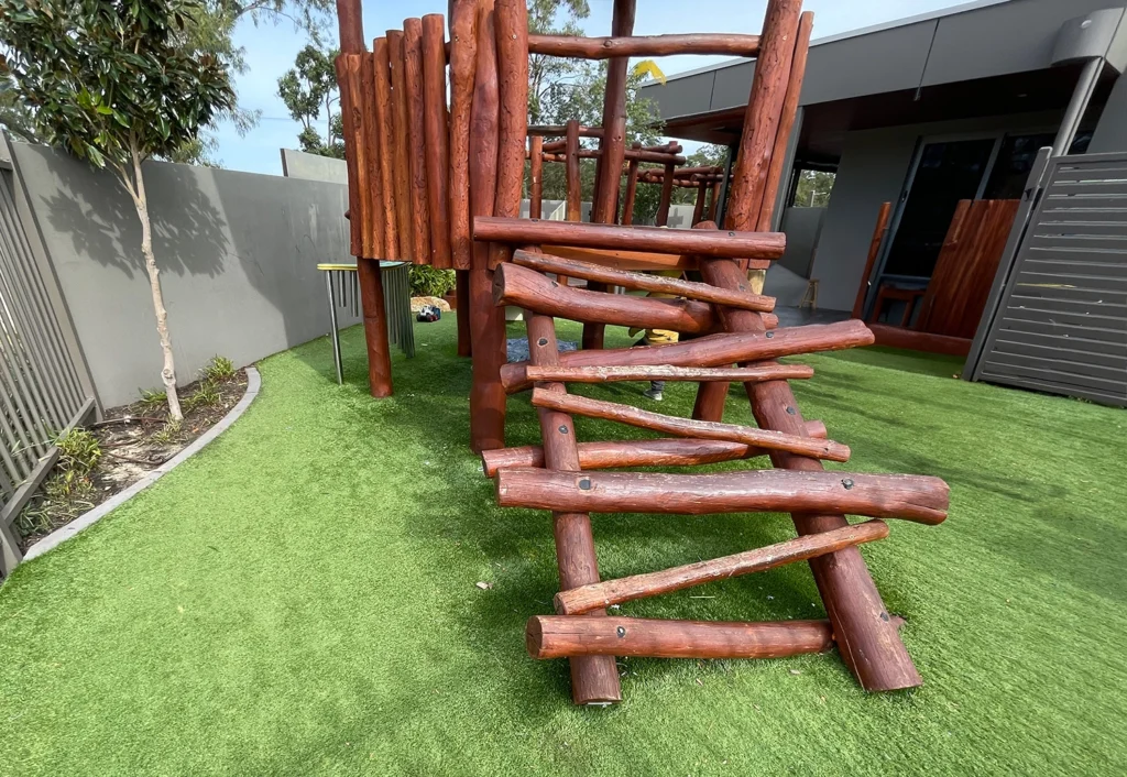 Wooden play structure with angled logs for children to climb on artificial soft fall grass next to a modern daycare and kindergarten building.