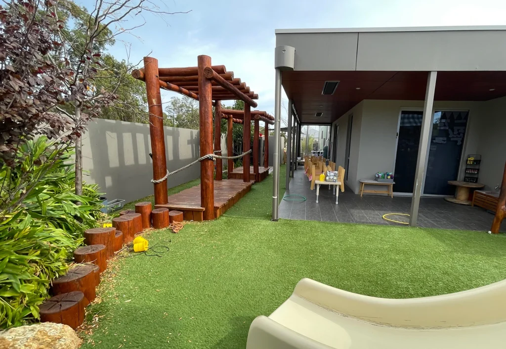 Outdoor play area at Busy Bees Byford South featuring a slide, wooden log stepping stones, and a shaded seating area.