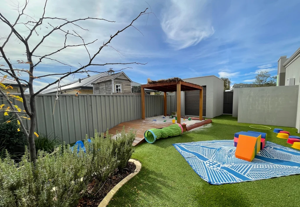 An outdoor play area at Byford childcare centre with a wooden deck, sandpit and green artificial turf with a fabric tunnel toy, a blue playmat and soft blocks.