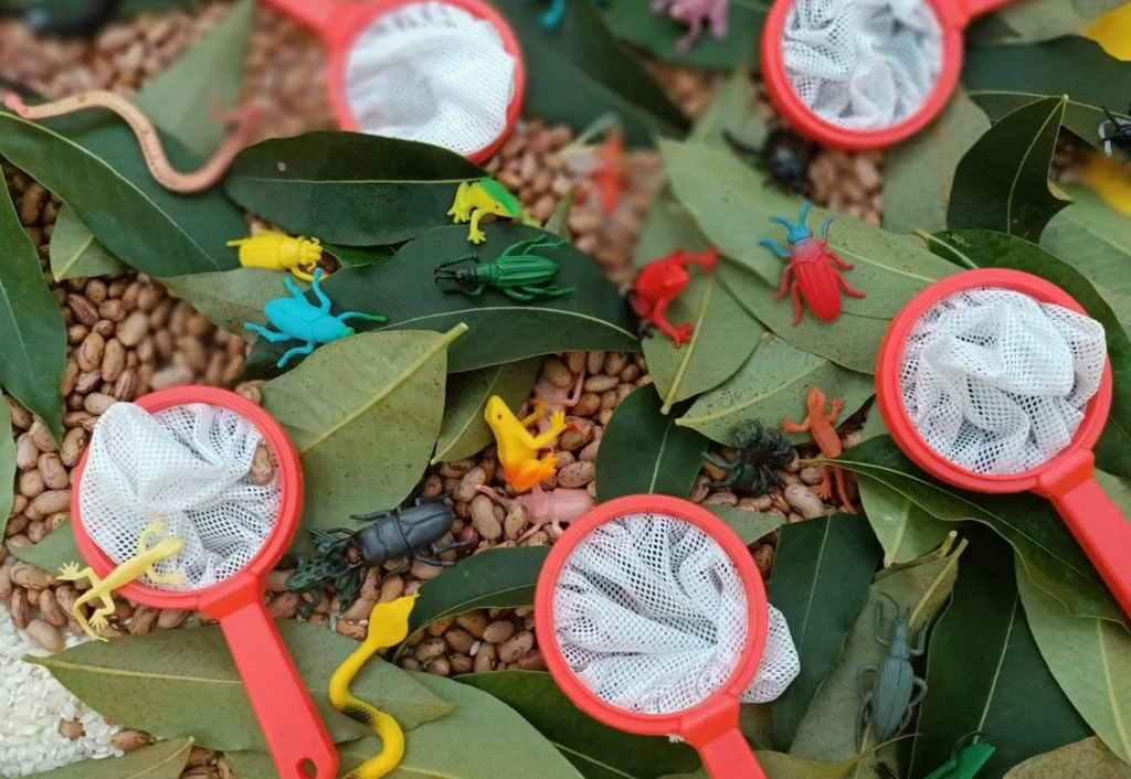 Toy insects, green leaves, red-handled mesh nets, and brown beans are scattered in this play scene, creating a colorful and interactive setting.