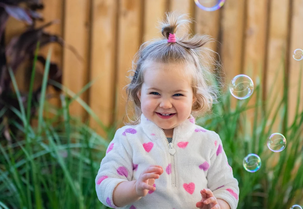 A toddler with a top-ponytail, wearing a white jacket with pink hearts, smiles while playing with bubbles in front of a wooden fence and greenery.