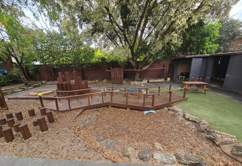 A playground with wooden structures, a bridge, stepping logs, and a picnic table. There are trees and foliage surrounding the area and a building in the background.