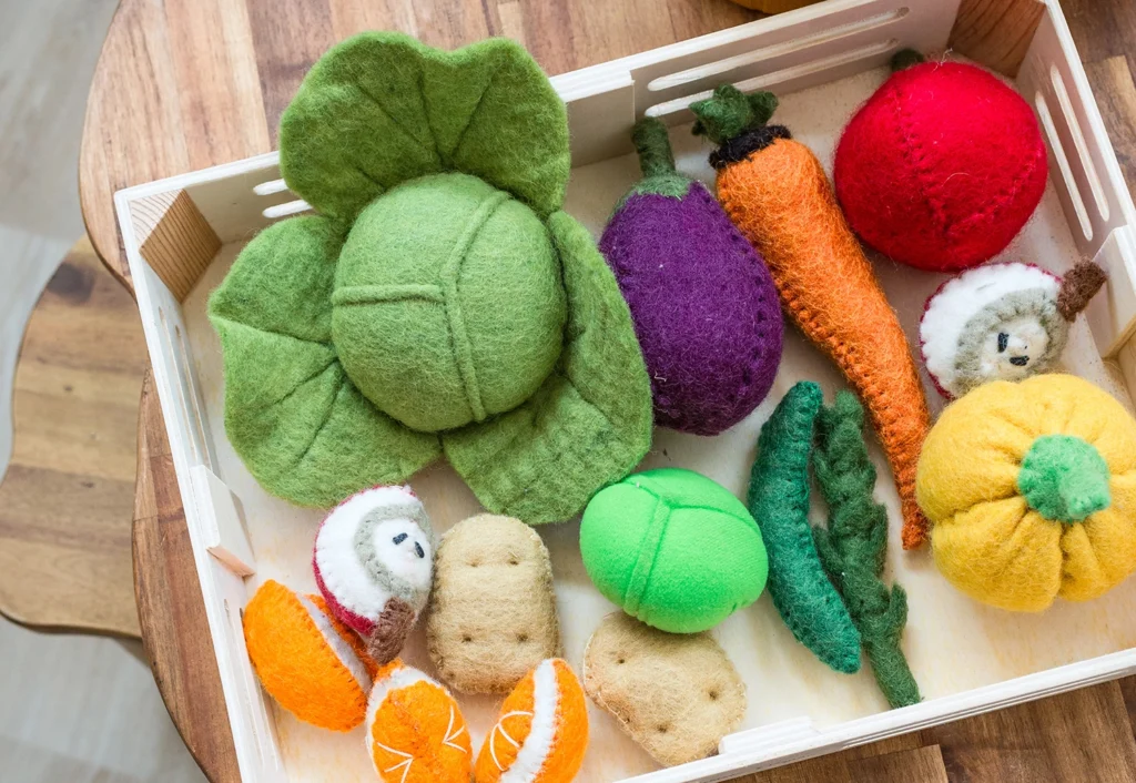 A wooden tray contains various handmade felt vegetables and fruits, including a cabbage, eggplant, carrot, radish, pumpkin, orange slices, and potatoes.