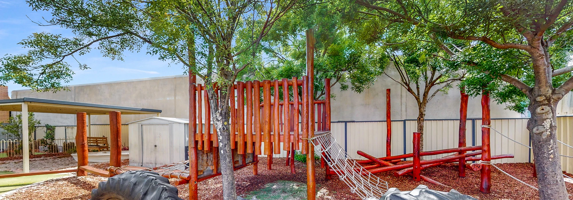 Outdoor play area with wooden climbing structures, a rope bridge, tire obstacles, and shaded spaces, surrounded by trees and a fence.