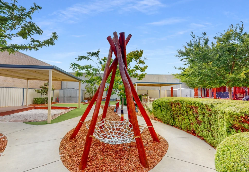 Playground area featuring a red wooden teepee structure with a net hammock, surrounded by pathways, vegetation, and a sandy play area under a canopy.