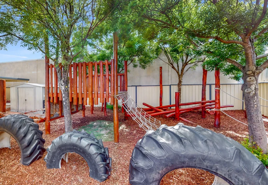 Outdoor playground with large tractor tires, wooden climbing structures, rope bridges, and surrounding trees. Mulch covers the ground, and there is a white shed in the background.