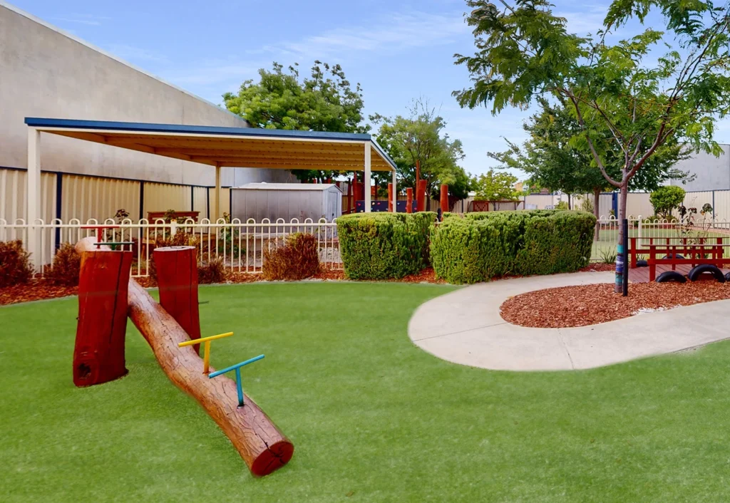 A playground with a seesaw, green grass, a white picket fence, and a shaded area is surrounded by trees and bushes. A concrete path curves through the lawn.