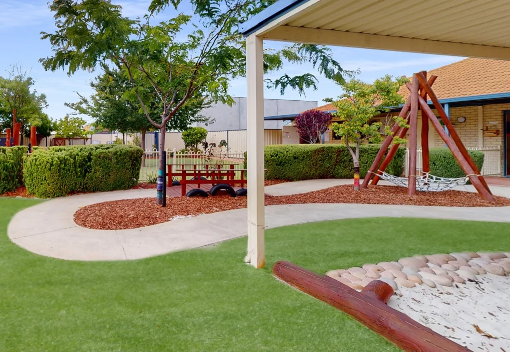 A playground area with green grass, a small bridge, swings, and a covered area. Pathways run through the well-maintained landscape with trees and bushes.