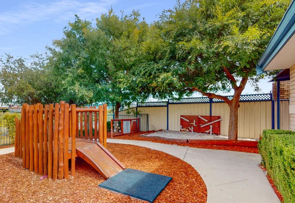 Outdoor playground area with a small wooden slide, surrounded by mulch, trees, and a white fence with a red-paint design. There is a paved walkway and shrubs near a building.