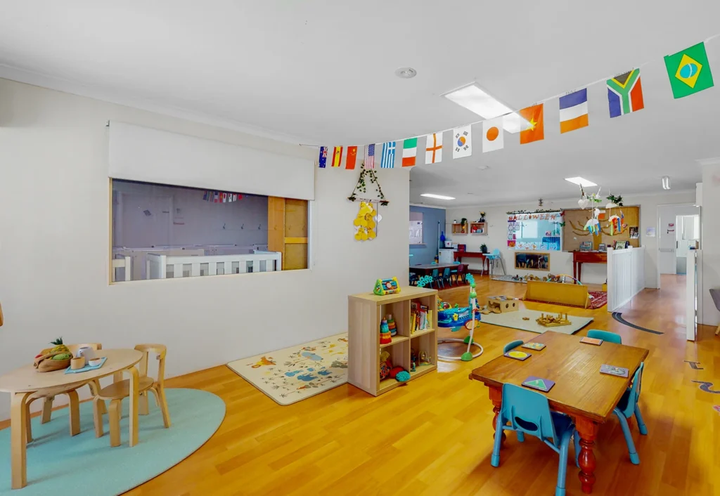 A bright, spacious classroom with international flags hanging from the ceiling, wooden furniture, a small play area with a mat, toys, books, and colorful decorations.