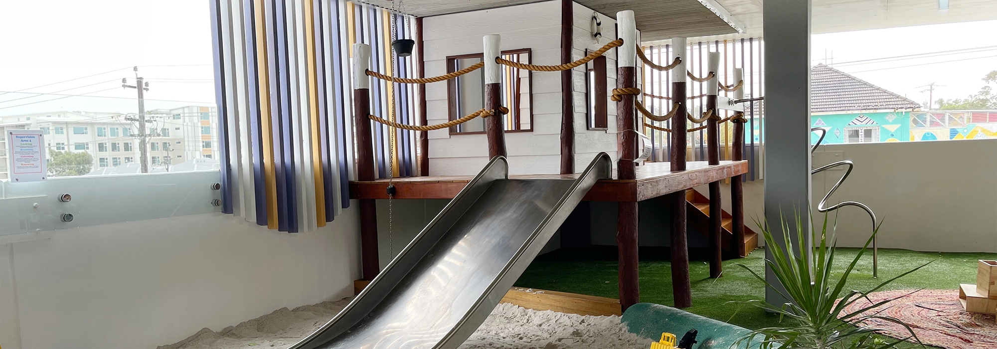 A playground slide attached to a structure resembling a ship's deck with rope railings, located indoors with artificial grass and sand surrounding the area.