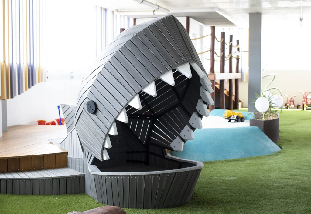 Wooden play structure shaped like a shark's open mouth in an indoor playground with green turf and various play equipment in the background.