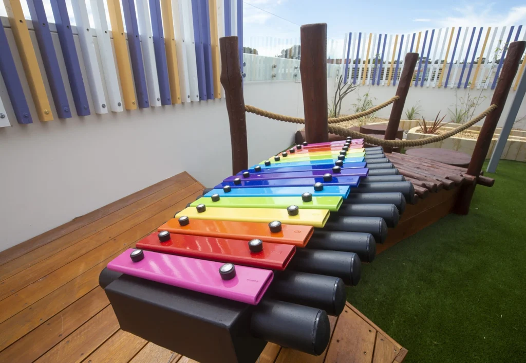 A colorful outdoor xylophone with rainbow-colored keys is set beside a wooden walkway on artificial turf, with a backdrop of vertical pastel-colored bars.