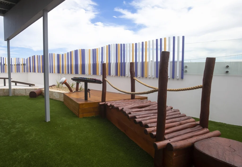 Playground area with a wooden bridge, rope handrails, and artificial grass. Colorful vertical poles serve as a decorative fence in the background under a partially cloudy sky.