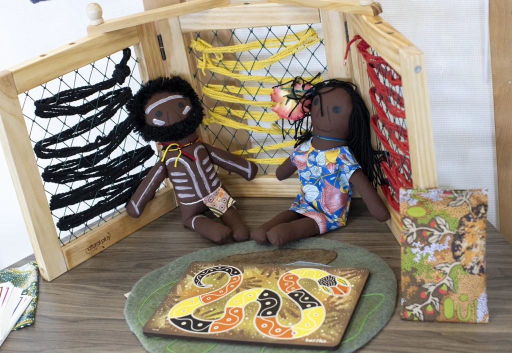 Two fabric dolls with brown skin tones and traditional attire sit within a wooden display with colorful strings and indigenous art patterns. A snake-themed art piece is also on the table.