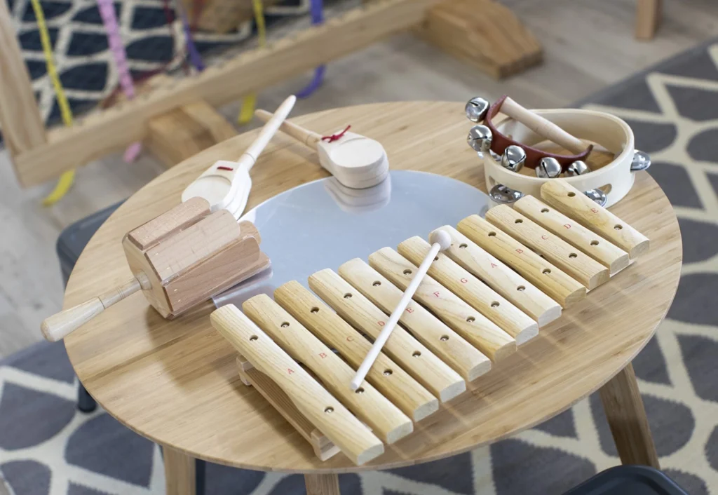 A round wooden table displays a xylophone with mallets, a tambourine with jingles, and a wooden percussion instrument. The background includes a wooden frame and geometric patterned carpet.