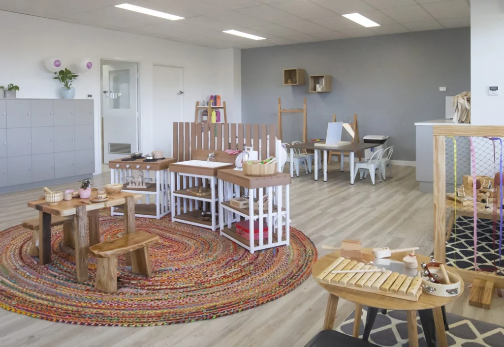 A classroom with wooden tables, chairs, and play structures on a colorful circular rug. The walls are grey and white, with cabinets, shelves, and children's art supplies displayed.