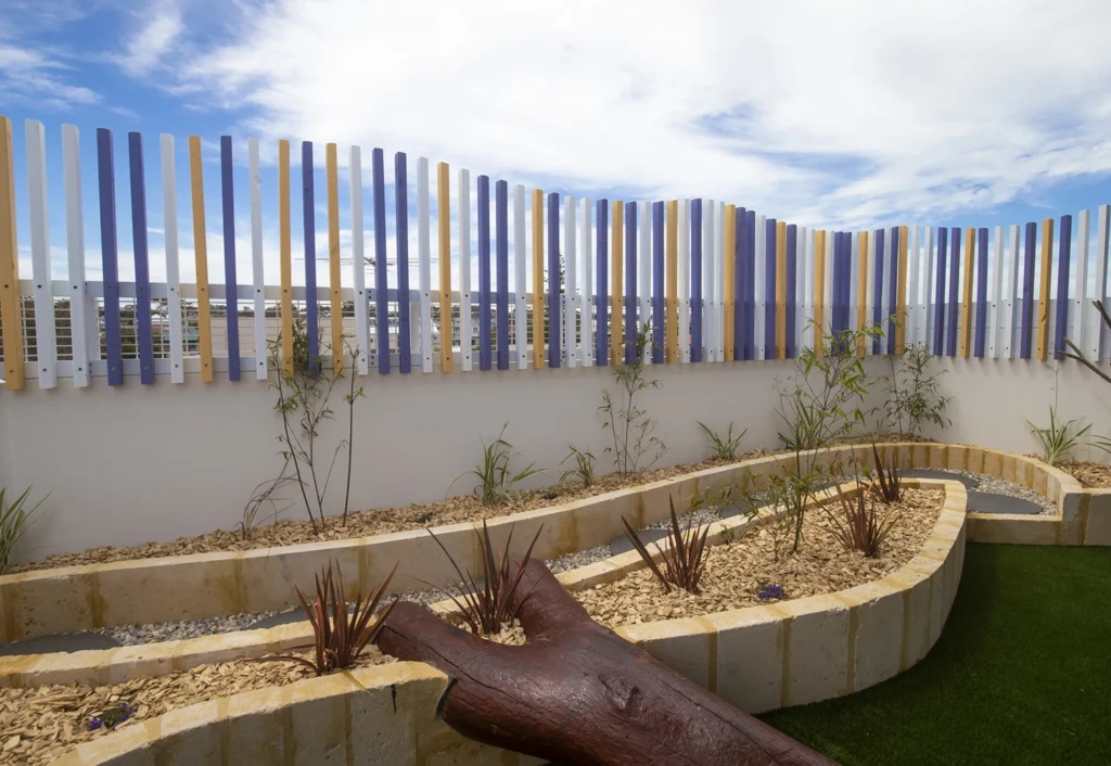 A curved, tiered garden bed with small plants is bordered by a low wall. Vertical blue, white, and yellow fence panels are in the background under a partly cloudy sky.