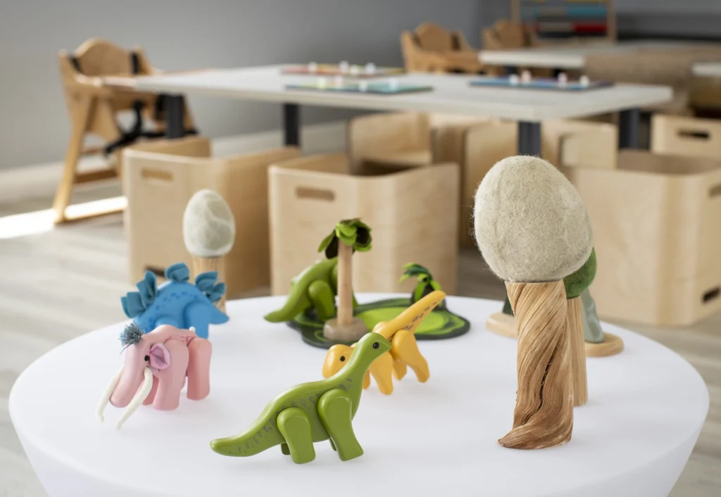 A round table with a collection of colorful wooden toy animals, including dinosaurs and an elephant, in front of a classroom setting with child-sized furniture and storage boxes.