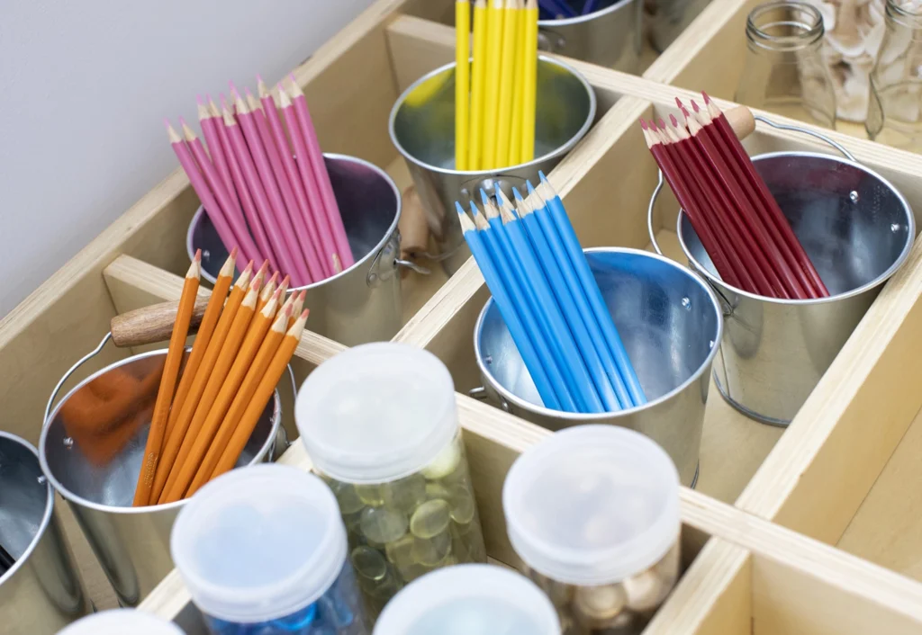 Metal buckets hold various colored pencils organized by color, set within a wooden grid. Clear containers of craft supplies are also visible.