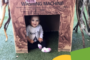 A baby, sitting inside a cardboard box labeled "Washing Machine," smiles while looking at the camera. The box appears to be part of a play area or early learning outdoor setting.