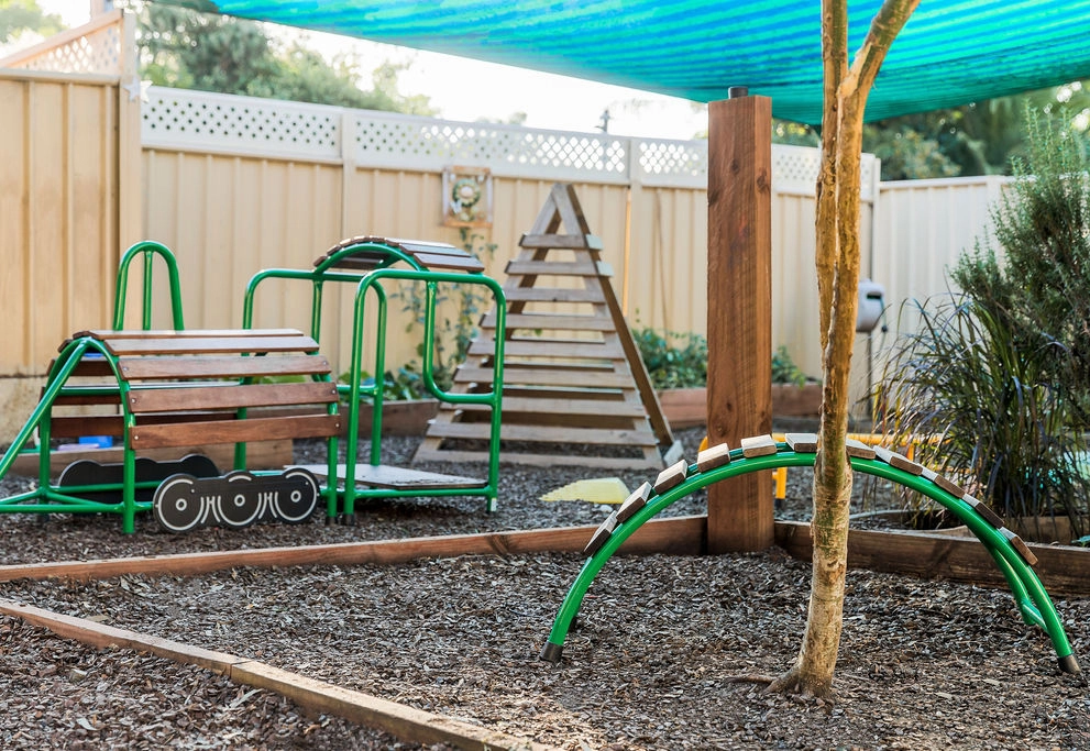 A fenced playground featuring various outdoor play equipment, including a wooden train structure, a triangle climbing frame, and a green arch, with a blue shade cloth overhead.